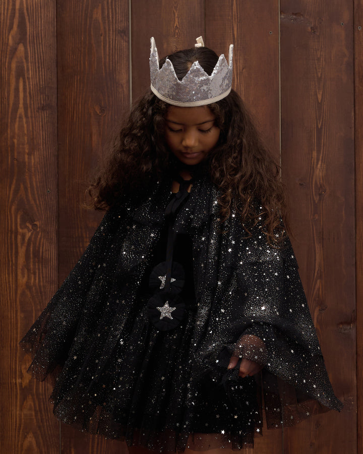 A young girl with long curly brown hair wearing a black sparkly cape with silver star details and a silver crown, standing against a dark wood plank background.