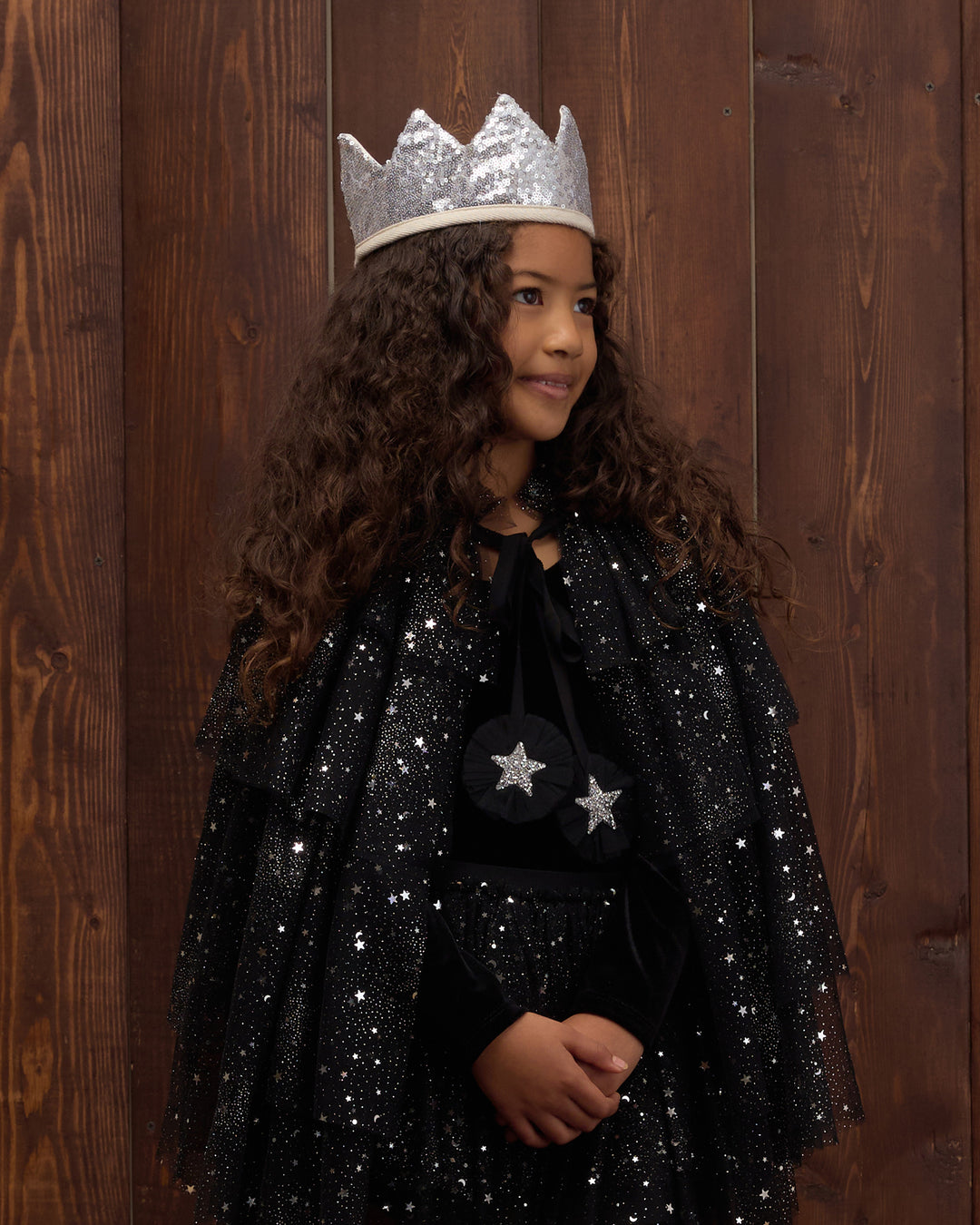 A young girl with long curly brown hair wearing a sparkly black cape with silver star details and a silver crown, standing against a dark wood plank background.