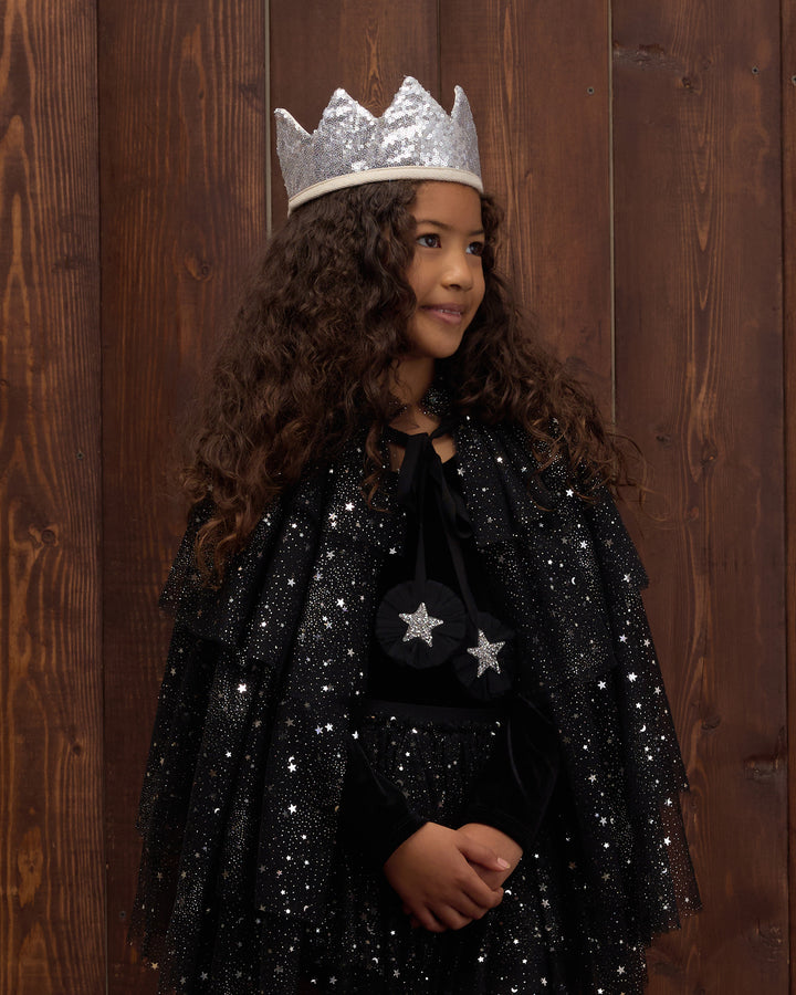 A young girl with long curly brown hair wearing a sparkly black cape with silver star details and a silver crown, standing against a dark wood plank background.
