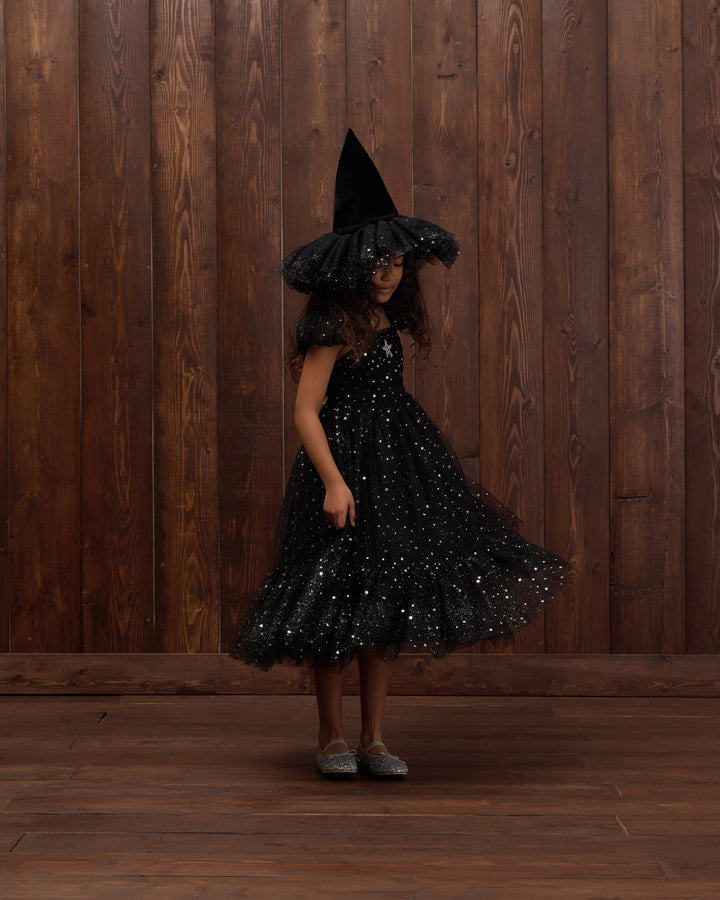 A young girl wearing a black witch costume with a tall pointed hat and a sparkly black dress, standing against a dark wood plank wall and floor.