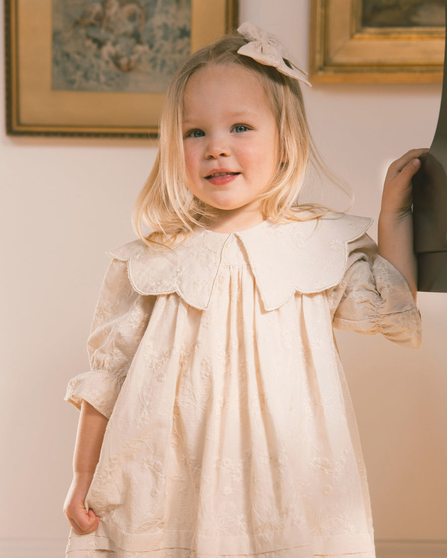 A young blonde girl wearing a cream-colored embroidered dress with a large ruffled Peter Pan collar and a matching bow in her hair, standing in an indoor setting.
