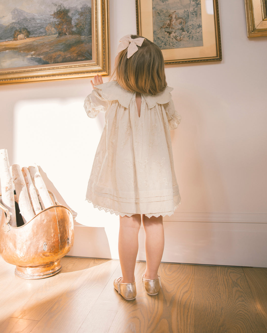 A young girl with a pink bow in her hair stands with her back to the camera, wearing a cream-colored lace dress and metallic gold shoes in a sunlit room with framed paintings and a copper pot.