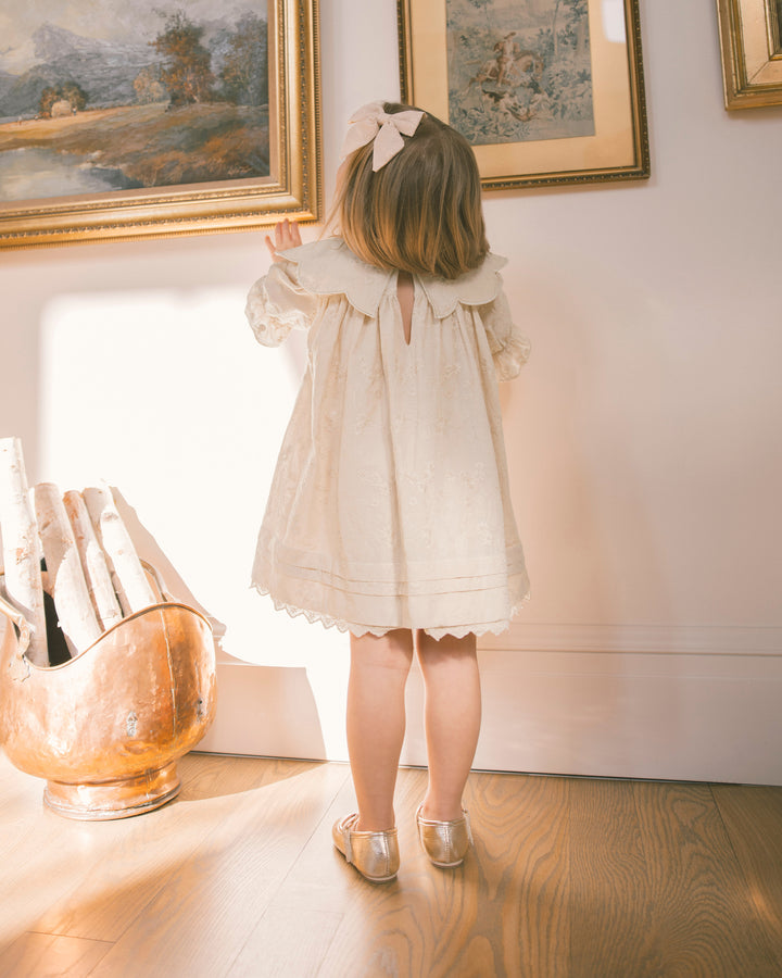 A young girl with a pink bow in her hair stands with her back to the camera, wearing a cream-colored lace dress and metallic gold shoes in a sunlit room with framed paintings and a copper pot.