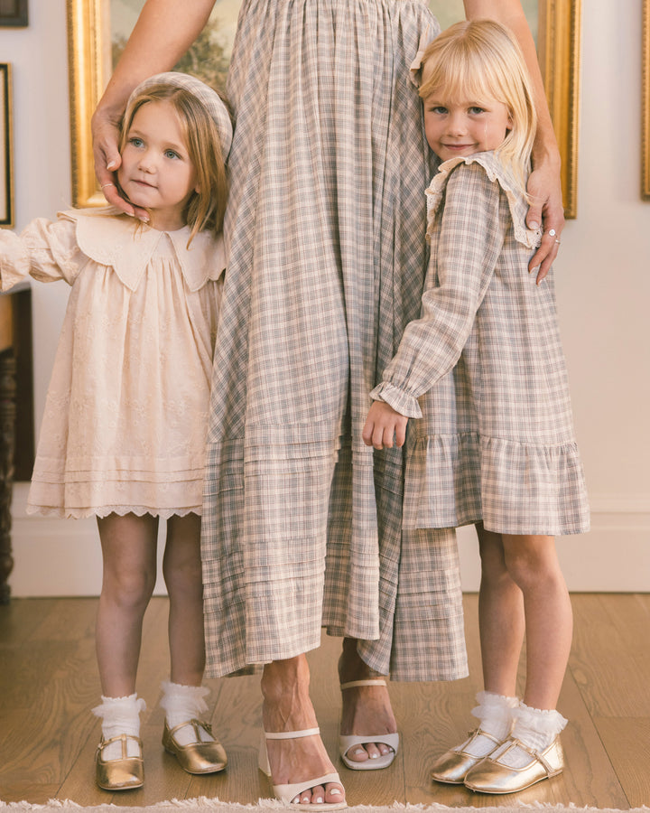 A mother and her two daughters wearing matching vintage-style plaid dresses in a warm, softly lit indoor setting. The mother wears a long beige and gray plaid dress, while the daughters wear matching shorter versions in different tones of the plaid pattern.