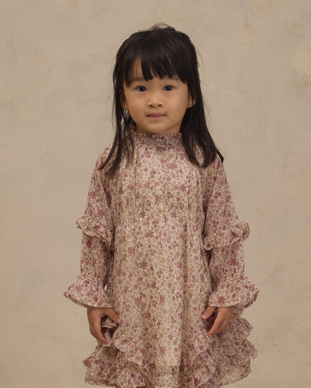 A young girl with dark hair wearing a long-sleeved, high-neck dress with a pink and beige floral pattern and ruffled details, standing against a textured beige background.