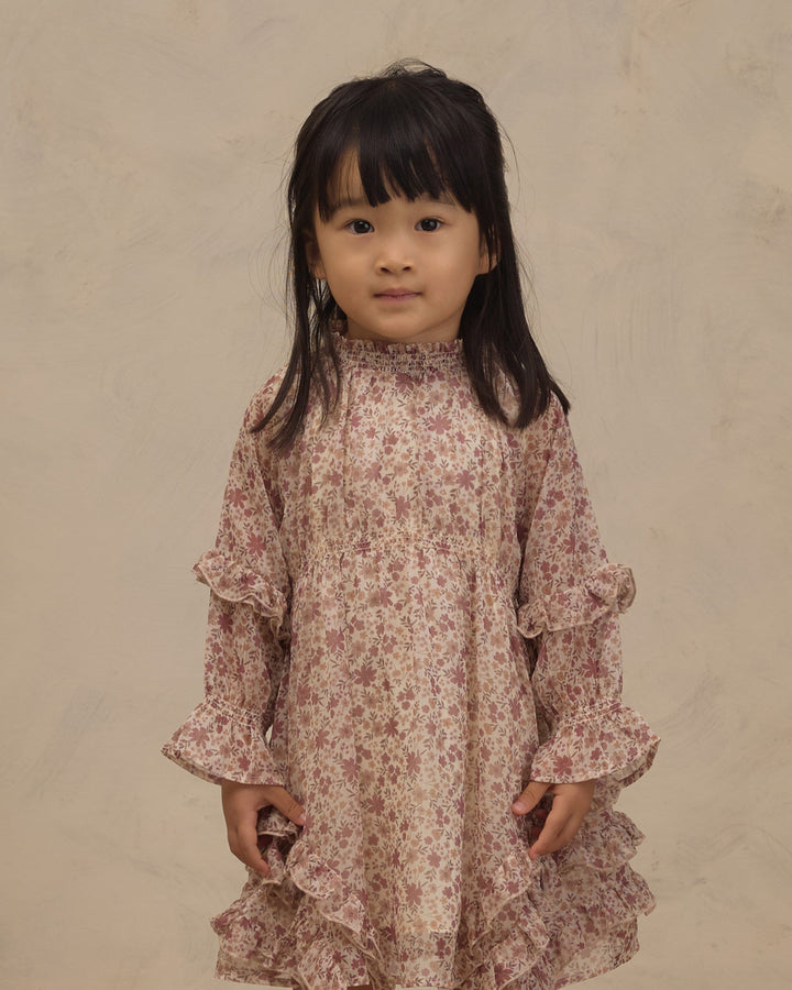 A young girl with dark hair wearing a long-sleeved, high-neck dress with a pink and beige floral pattern and ruffled details, standing against a textured beige background.