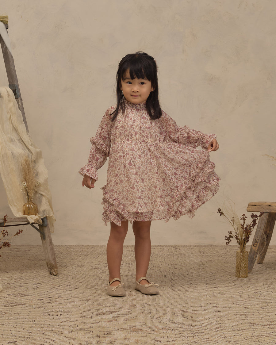 A young girl with dark hair stands in a studio wearing a long-sleeved, high-neck floral dress in a dusty rose and beige pattern. She is holding out the side of the dress to show its volume.
