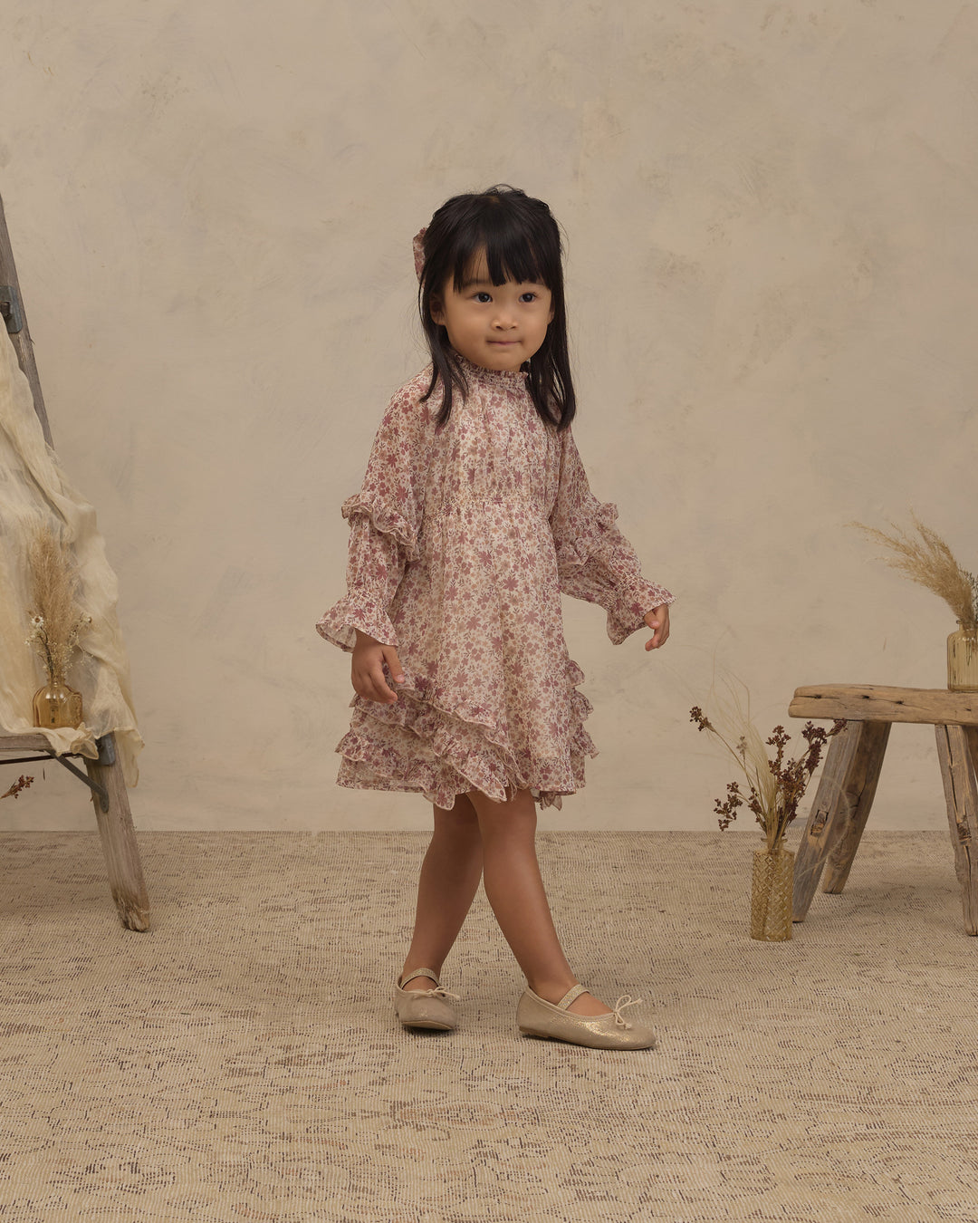 A young girl with dark hair stands in a studio setting wearing a long-sleeved, ruffled dress with a pink and beige floral pattern, paired with beige ballet flats.