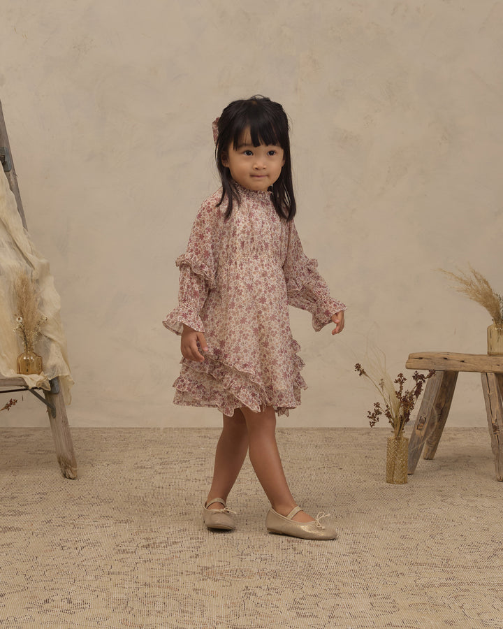 A young girl with dark hair stands in a studio setting wearing a long-sleeved, ruffled dress with a pink and beige floral pattern, paired with beige ballet flats.
