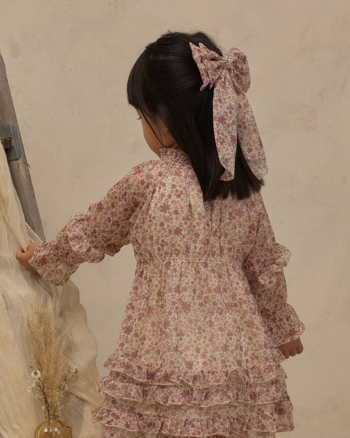 A young girl with long black hair wearing a pink and beige floral long-sleeve dress with a matching hair bow, photographed from behind against a textured beige wall.