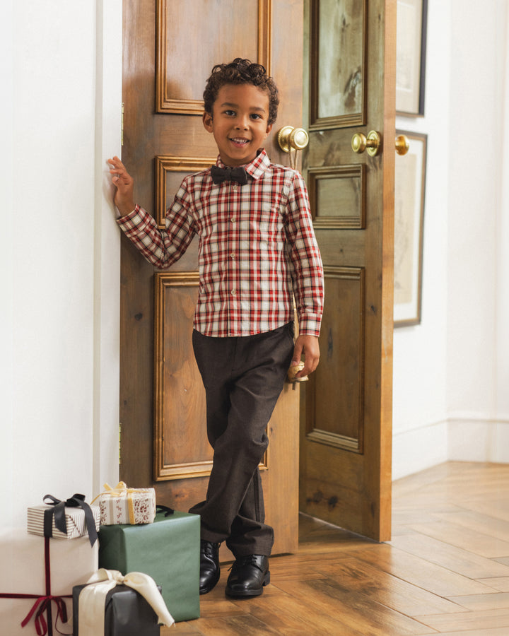 A young boy stands in a doorway wearing a red and white gingham long-sleeve shirt, a black bow tie, dark trousers, and black shoes. Gift boxes are stacked on the floor to his left.
