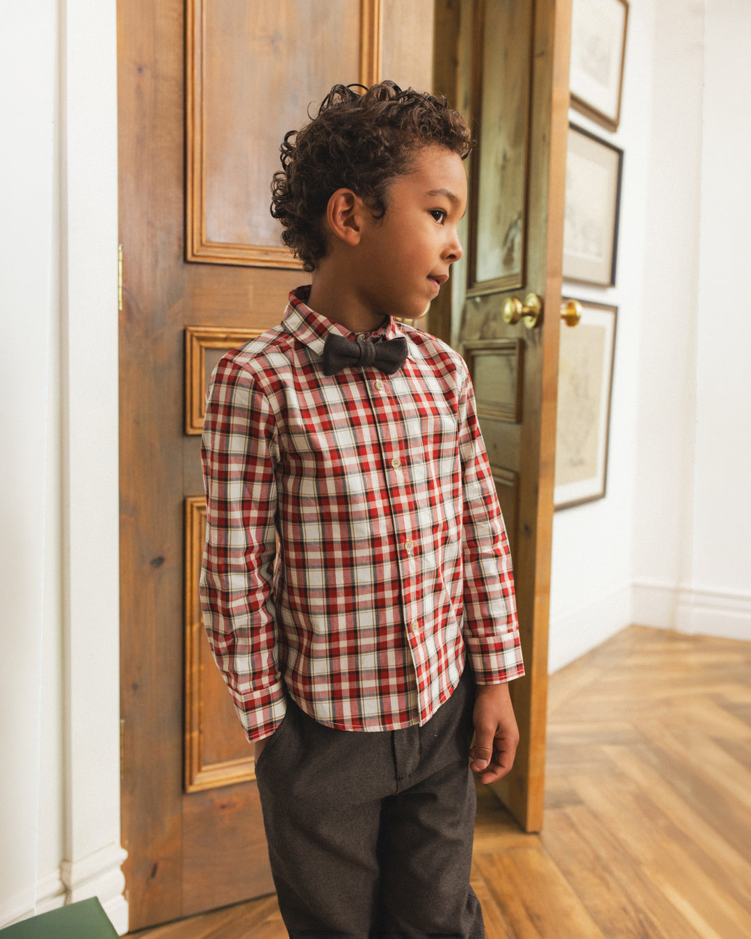 A young boy wearing a red and white plaid long-sleeve dress shirt with a black bow tie and dark gray trousers, standing in a doorway in a home interior.