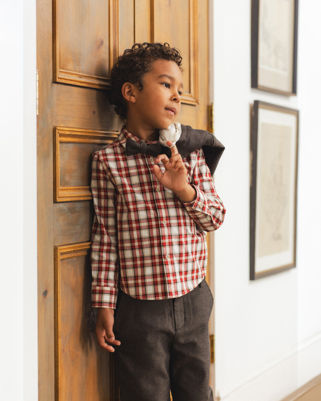 A young boy with curly dark hair stands leaning against a wooden door in a home interior. He is wearing a red, white, and beige plaid long-sleeve button-up shirt, dark gray trousers, and a dark gray bow tie with a white necktie peeking out.