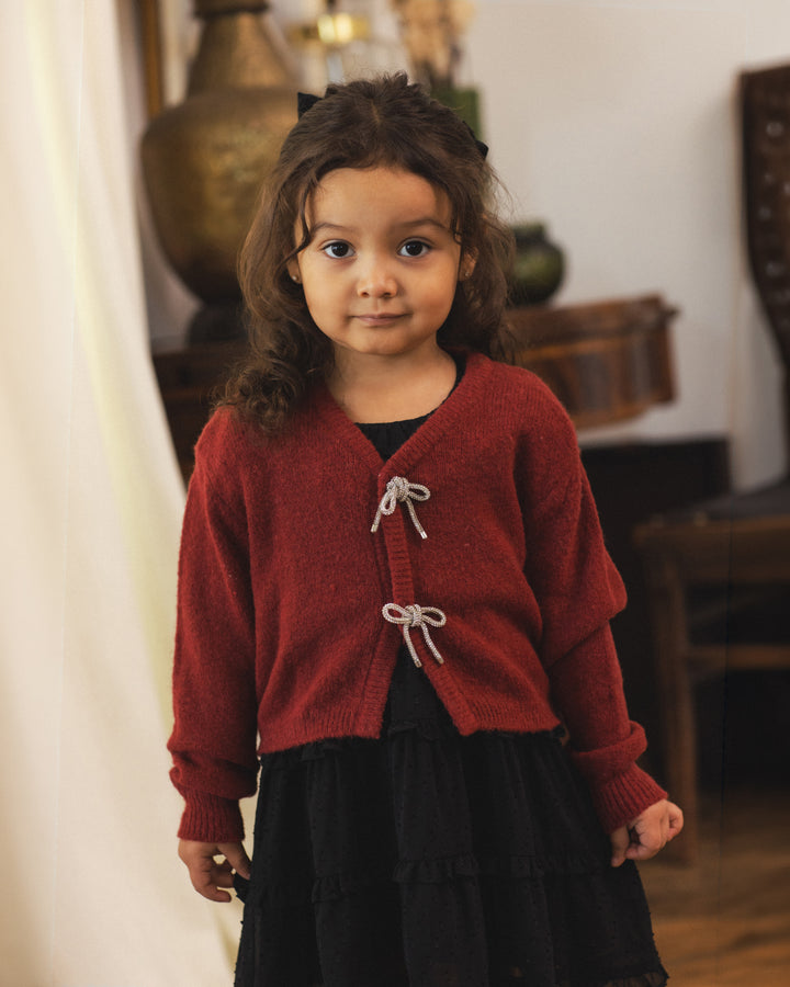 A young girl with curly brown hair wearing a red knit cardigan with white cord ties and a black dress, standing in a softly lit indoor room.