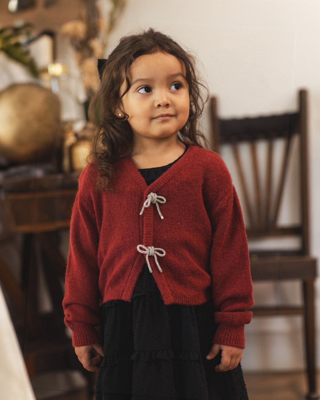A young girl with long brown hair wearing a red knit cardigan with white cord ties over a black outfit, standing in a softly blurred indoor setting with a wooden chair in the background.