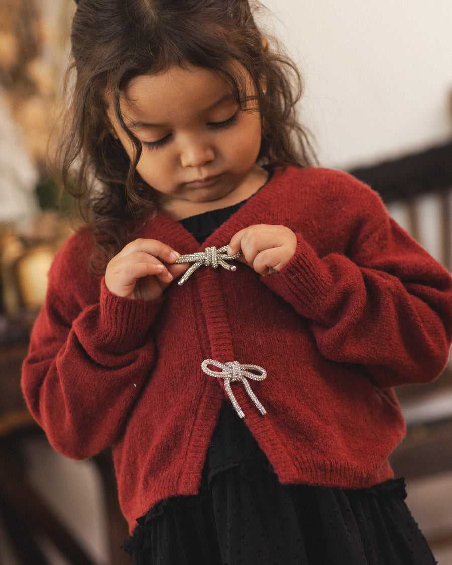 A young child wearing a red knit cardigan with silver bow details, standing in a softly blurred indoor setting.