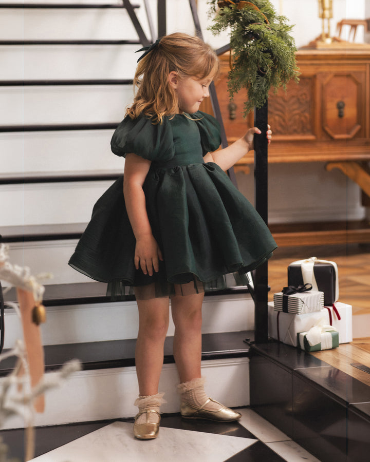 A young girl wearing a dark green puff-sleeve tulle dress and gold shoes stands on indoor stairs next to a decorated wreath and wrapped gifts.