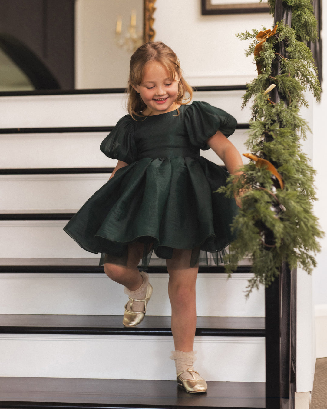 A young girl with blonde hair wearing a dark green puff-sleeve dress and gold shoes is smiling while walking down white stairs decorated with a green garland.