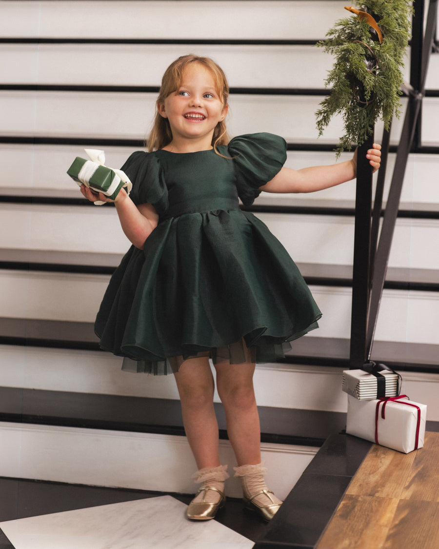 A young girl stands on a staircase wearing a dark forest green dress with puffed sleeves and a flared skirt. She is holding a wrapped gift in one hand and touching the stair railing with the other, with wrapped presents and a green garland nearby.