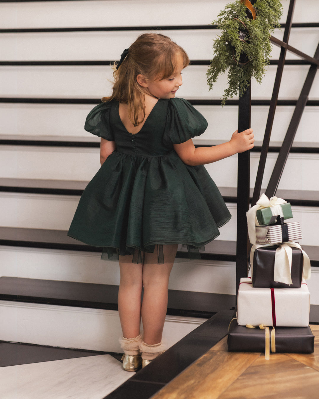 A young girl with light brown hair tied back wears a dark forest green puff-sleeve dress with a ruffled tulle skirt, standing on a black-and-white staircase next to a stack of wrapped Christmas presents.