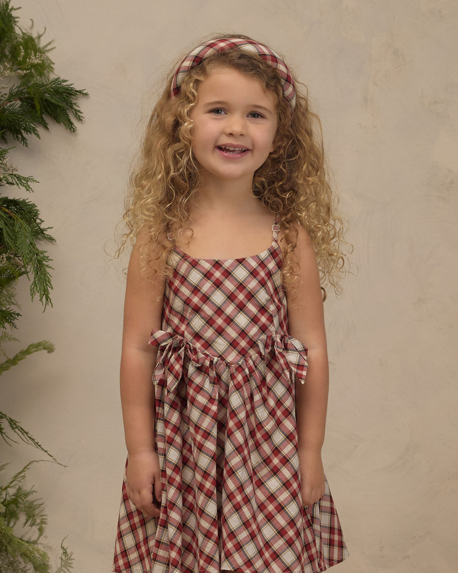 A young girl with curly blonde hair wearing a red and beige gingham dress with a matching headband, standing against a textured beige wall with green foliage on the left.