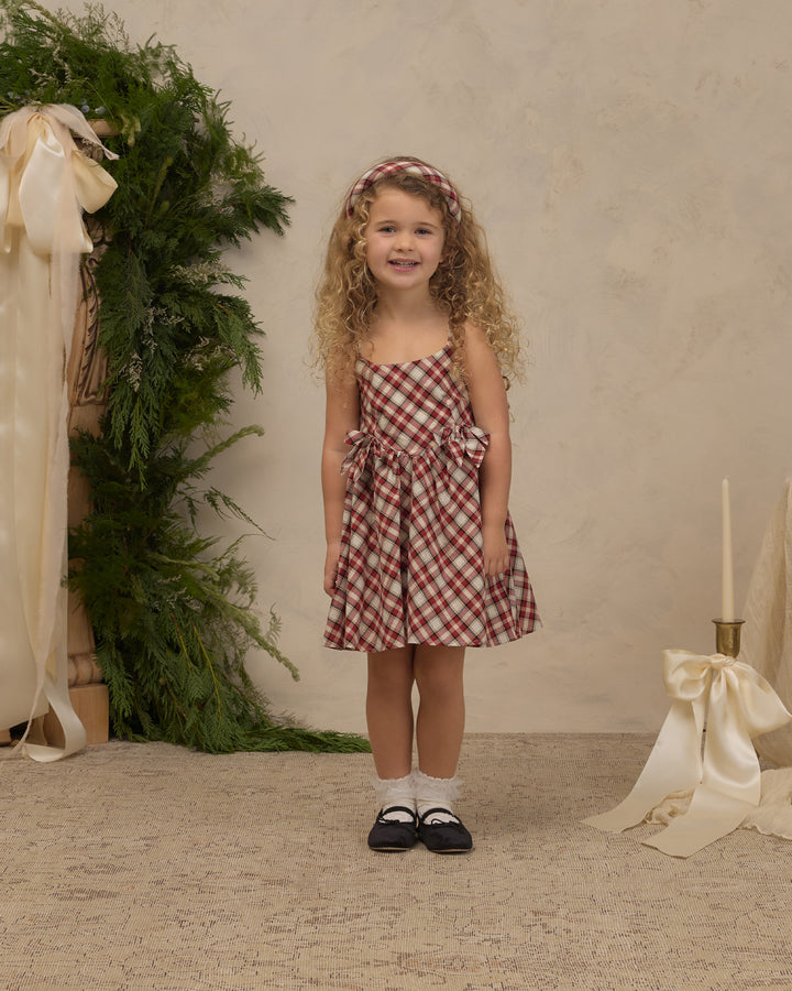 A young girl with curly blonde hair stands in a studio setting wearing a red and white gingham spaghetti strap dress with side bows, a matching gingham headband, white socks, and black Mary Jane shoes. The background features a textured beige wall, a green pine garland with a cream ribbon on the left, and a cream satin bow and candle holder on the right.