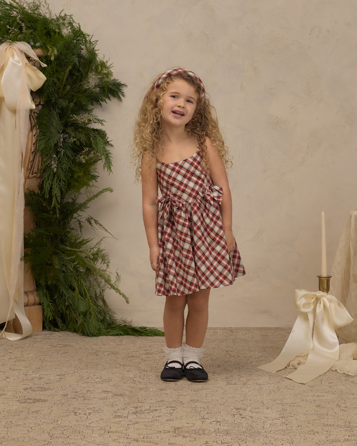 A young girl with curly blonde hair stands in a studio wearing a red and white gingham check spaghetti strap dress with a matching headband, white socks, and black Mary Jane shoes. The background features a textured beige wall, a green foliage arrangement on the left, and a cream ribbon and candle prop on the right.