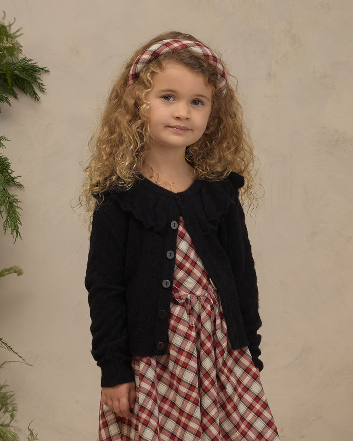 A young girl with curly blonde hair wearing a red and white plaid dress, a black knit cardigan, and a matching red and white plaid headband, standing against a textured beige wall.