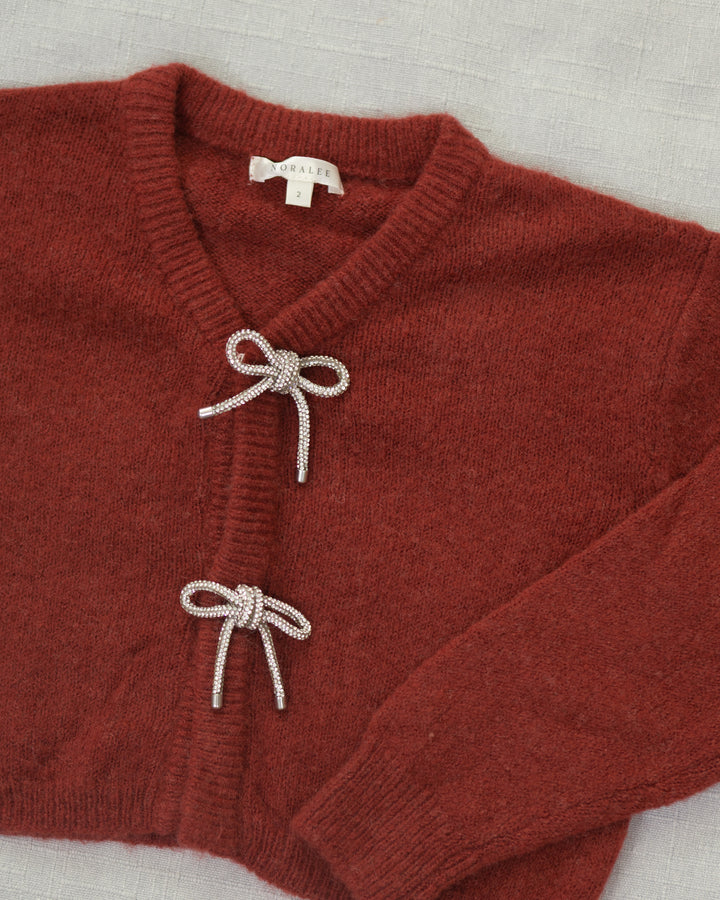 A close-up of a rust-red knit cardigan sweater with two decorative knotted bow details on the front, laid flat on a light textured fabric background. The brand name 'Noralee' is visible on the inner neck label.