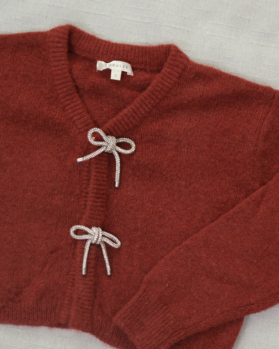 A close-up of a rust-red knit cardigan sweater with two decorative knotted bow details on the front, laid flat on a light textured fabric background. The brand name 'Noralee' is visible on the inner neck label.