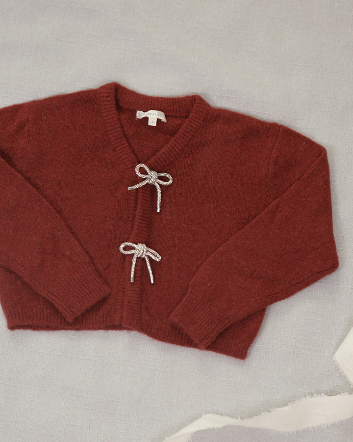 A rust-red knit cardigan sweater with white cord bow details on the front, laid flat on a light gray fabric background.
