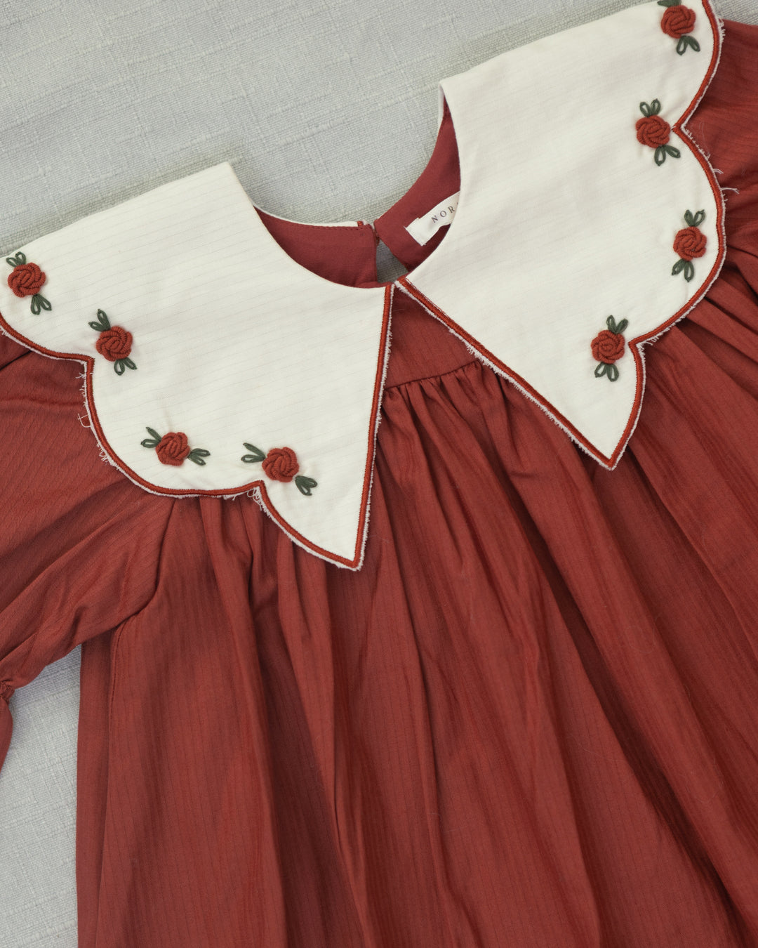 A close-up of a rust red dress featuring a large off-white Peter Pan collar decorated with embroidered red flowers and green leaves, laid flat on a light gray textured surface.