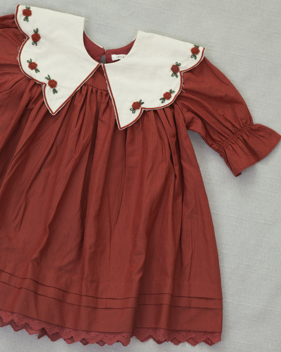A vintage-style children's dress in rust red with a large white Peter Pan collar decorated with red rose embroidery, laid flat on a gray textured background.