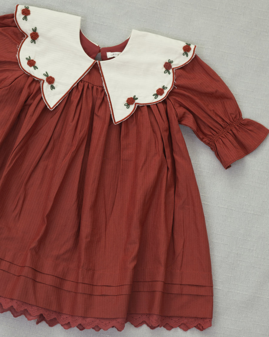 A vintage-style children's dress in rust red with a large white Peter Pan collar decorated with red rose embroidery, laid flat on a gray textured background.