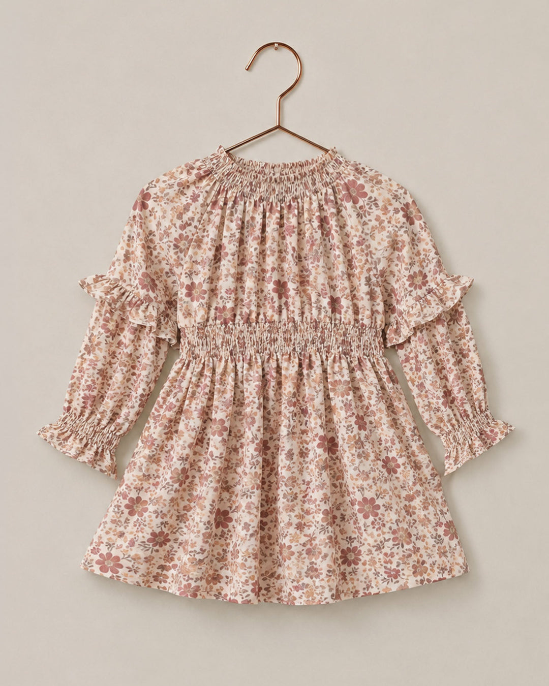 A long-sleeve child's dress with a delicate pink and brown floral print, featuring a smocked waist and ruffled sleeves, hanging on a wooden hanger against a solid beige background.
