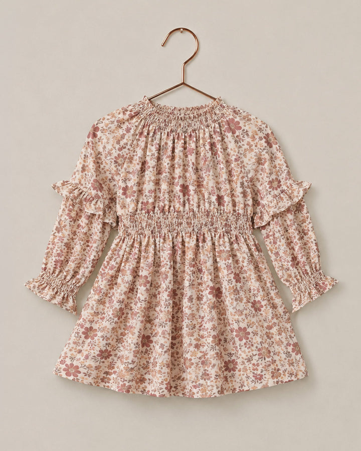 A long-sleeve child's dress with a delicate pink and brown floral print, featuring a smocked waist and ruffled sleeves, hanging on a wooden hanger against a solid beige background.