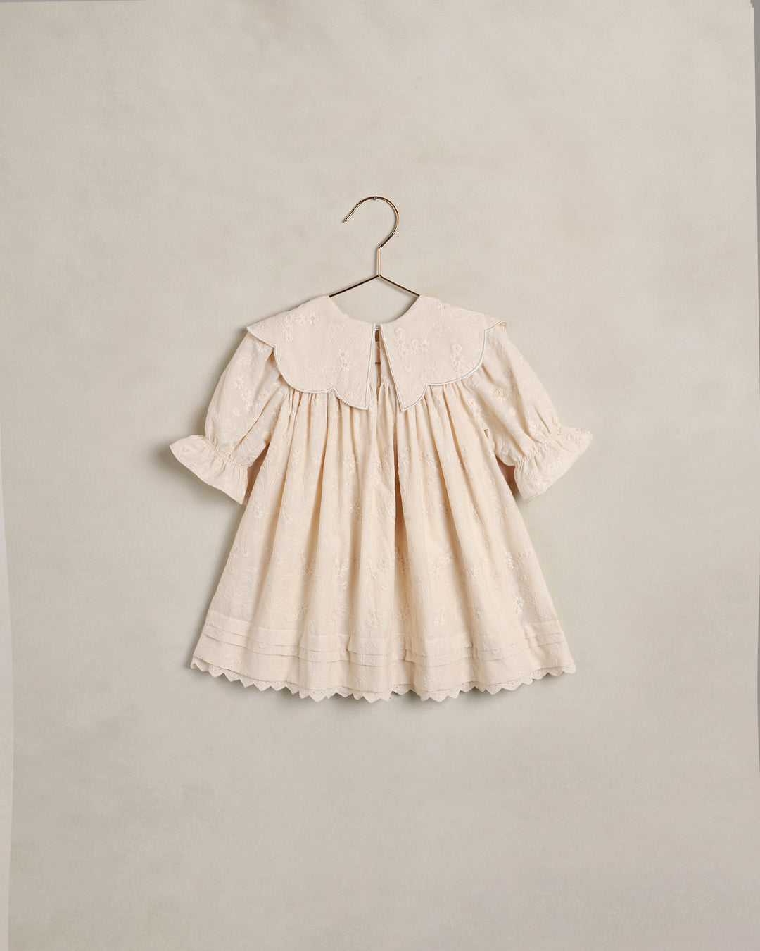 A cream-colored baby dress with a large embroidered Peter Pan collar, short puff sleeves, and a scalloped hem, hanging on a metal hanger against a textured beige wall.