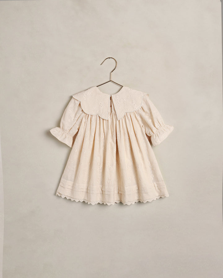 A cream-colored baby dress with a large embroidered Peter Pan collar, short puff sleeves, and a scalloped hem, hanging on a metal hanger against a textured beige wall.