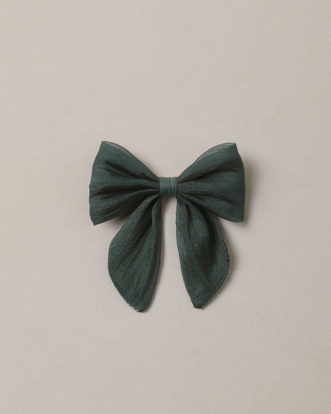 A dark green fabric hair bow with long tails, centered on a plain, warm off-white background.