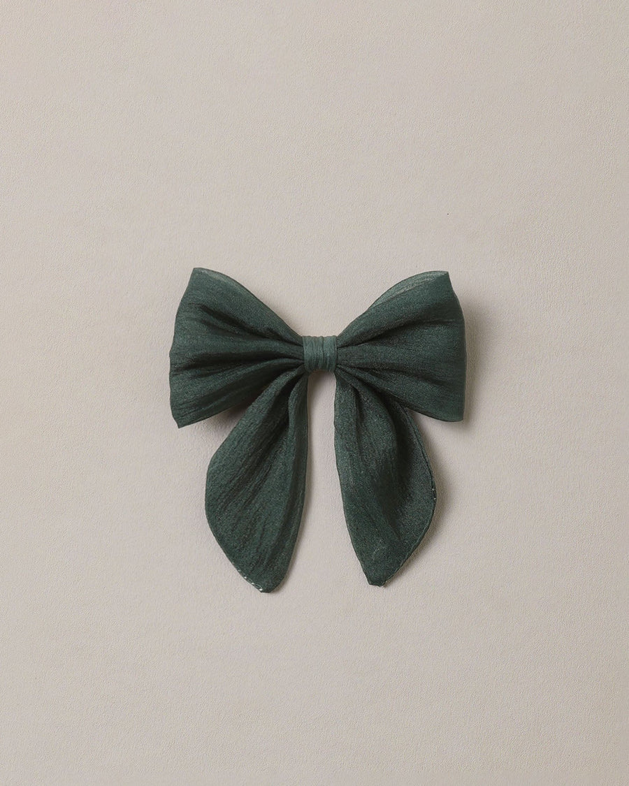 A dark green fabric hair bow with long tails, centered on a plain, warm off-white background.