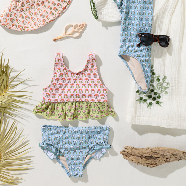 Children's floral swimsuits and accessories on a light background with palm leaves and sunglasses.