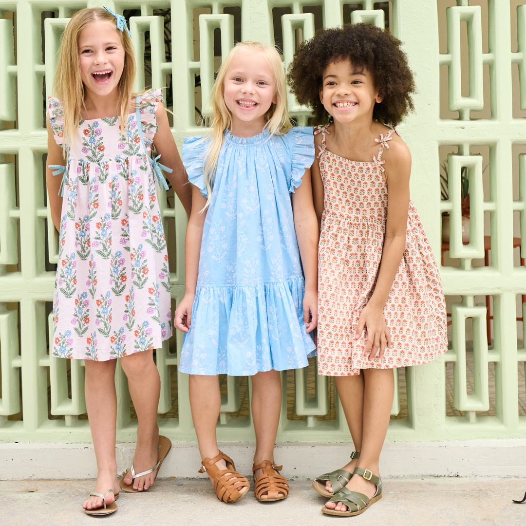 Three young girls wearing patterned dresses standing in front of a decorative wall.