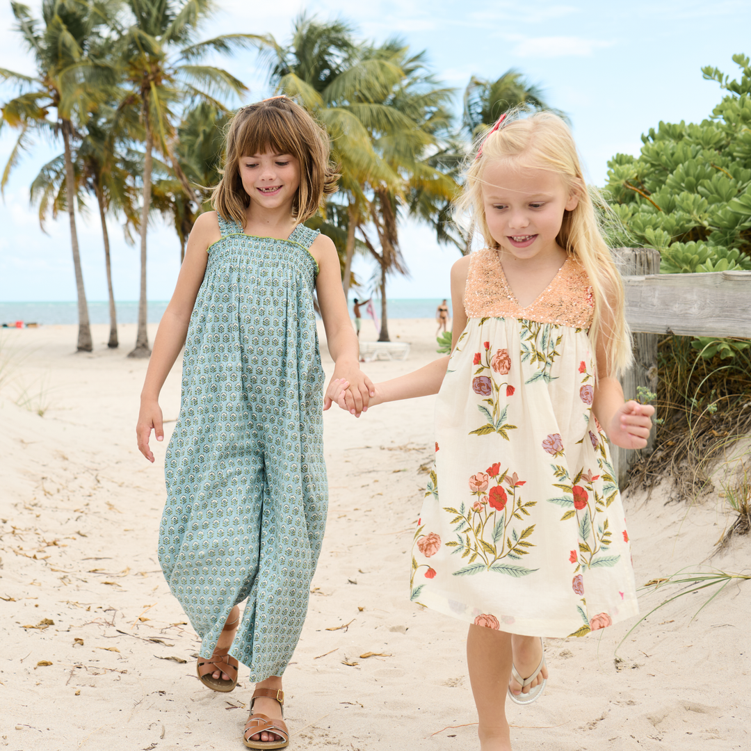 Two young girls holding hands on a sandy beach with palm trees in the background.
