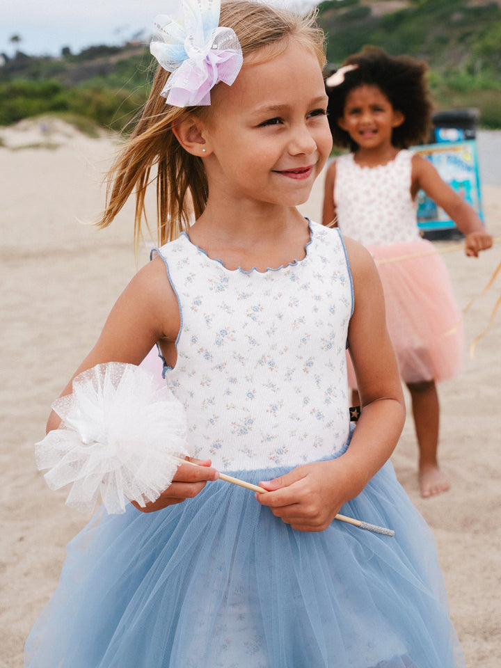 Young girl in a white top and blue tutu on a sandy beach with another child in the background.