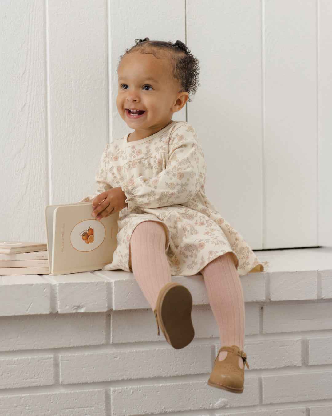 A smiling toddler girl sits on a white brick ledge holding a small beige book. She is wearing a cream long-sleeve dress with a delicate floral pattern, pink tights, and tan Mary Jane shoes against a white paneled wall background.