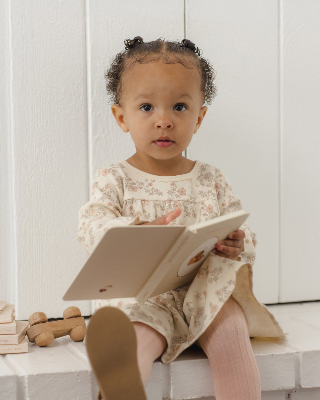 A toddler girl with braided hair sits on a white bench reading a book, wearing a cream floral long-sleeve dress and pink tights.