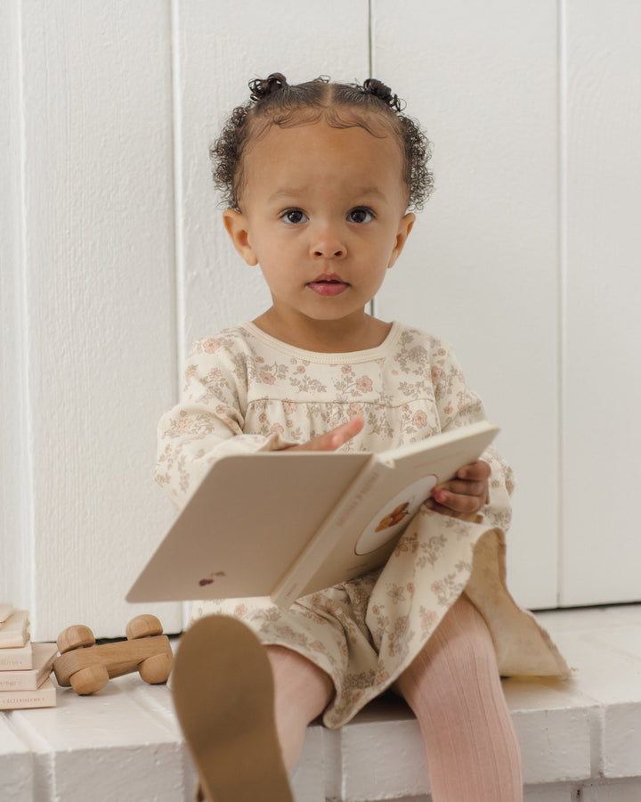 A toddler girl with braided hair sits on a white bench reading a book, wearing a cream floral long-sleeve dress and pink tights.