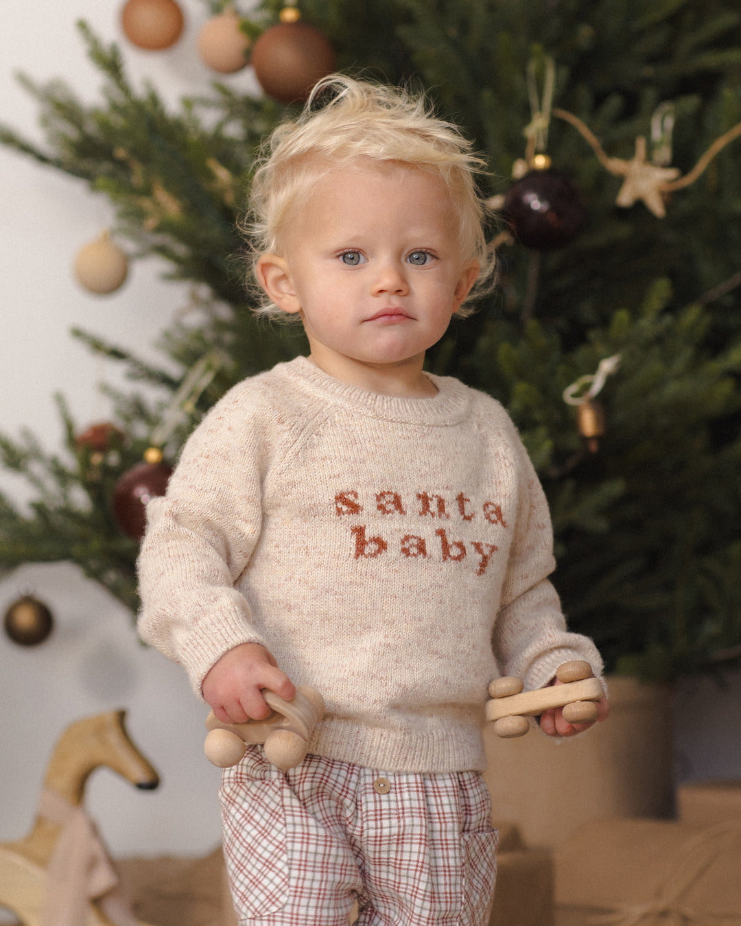 A toddler stands in front of a decorated Christmas tree wearing a cream knit sweater with 'santa baby' embroidered in brown across the chest, paired with plaid pants and holding wooden toy cars.