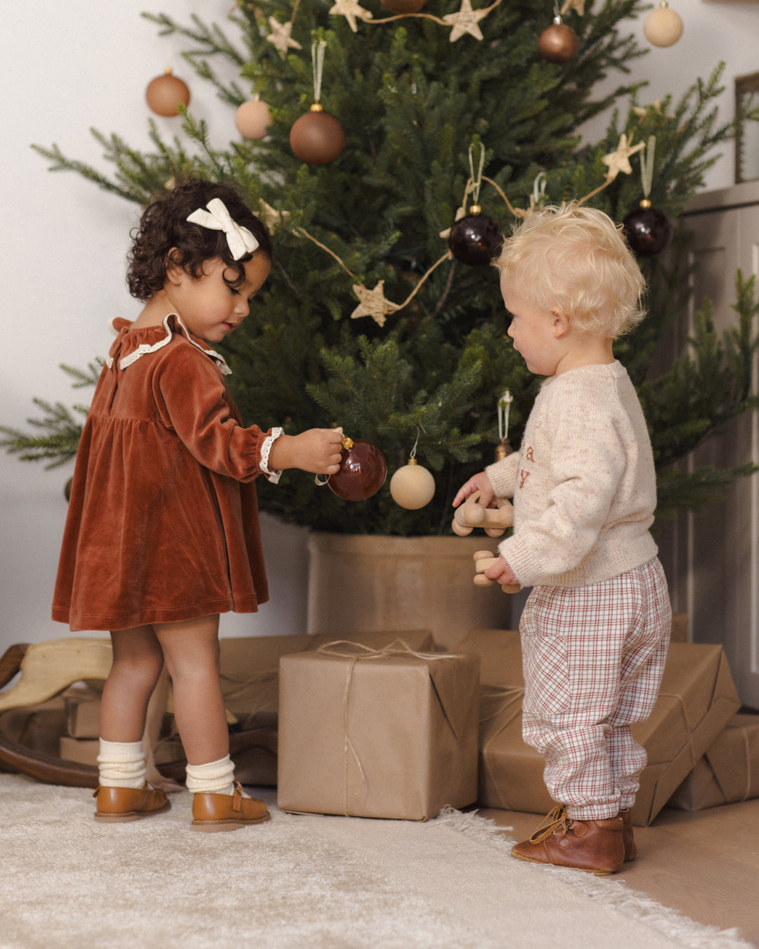 A toddler in a rust-brown velvet dress with a white bow and socks stands next to a baby in a cream sweater and plaid pants, decorating a Christmas tree with wooden ornaments. Wrapped gifts sit on the floor in front of the tree.