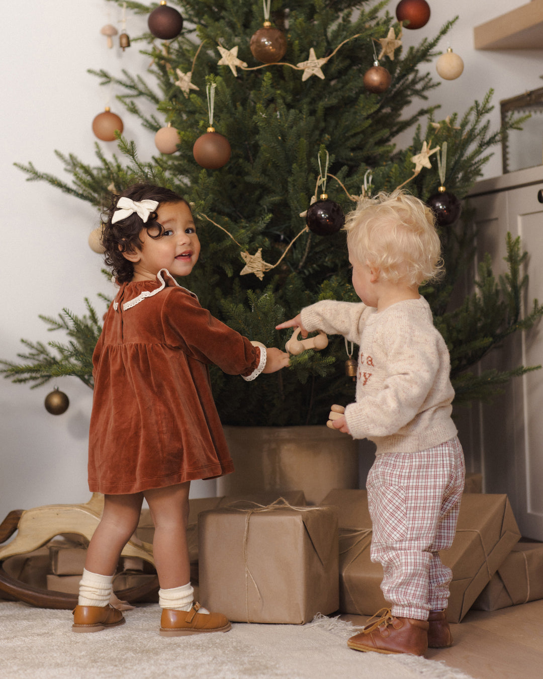 Two young children standing in front of a decorated Christmas tree, reaching toward each other. One child wears a rust-brown velvet dress with white trim, white socks, and brown shoes, while the other wears a cream sweater and plaid pants.